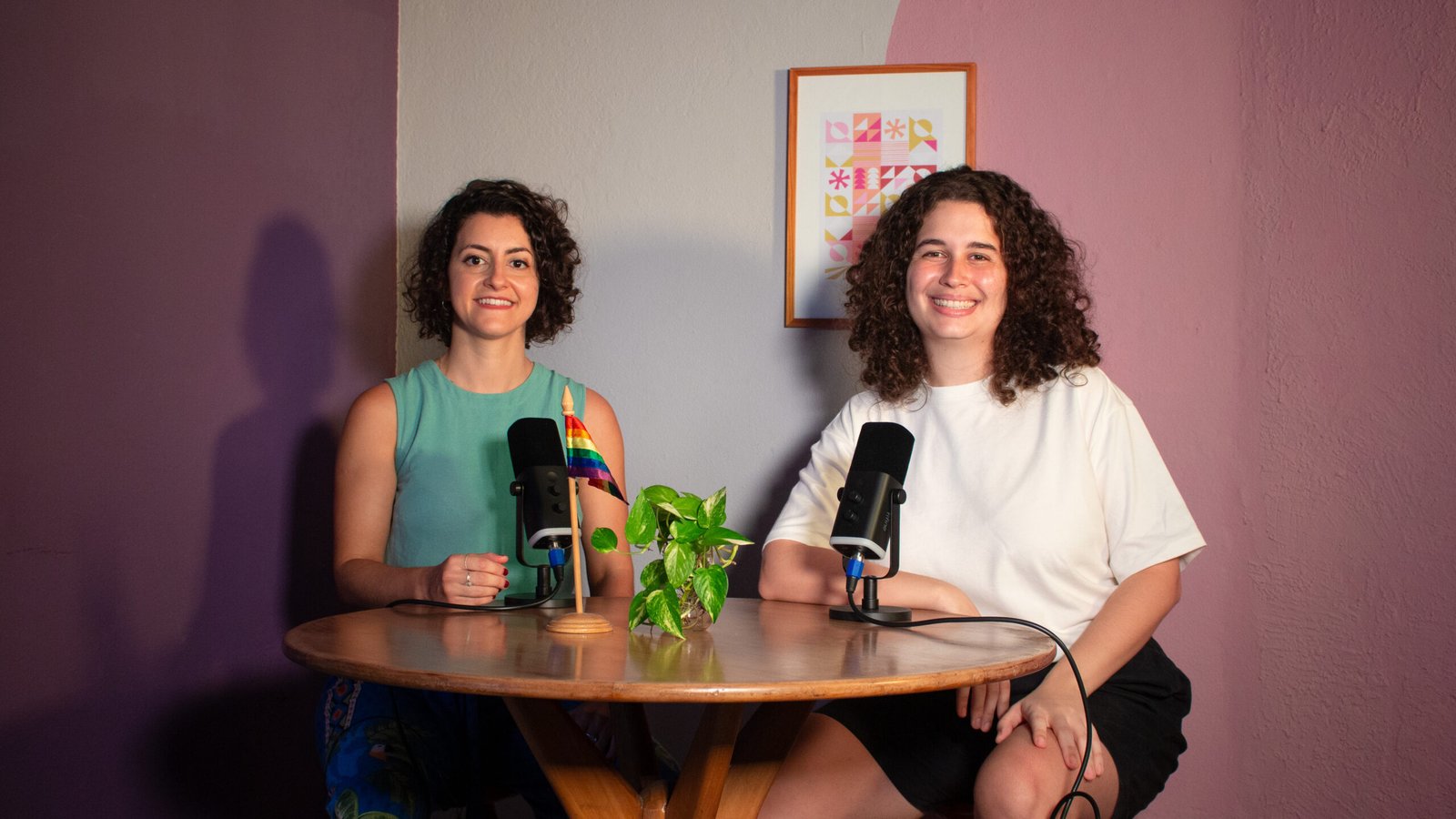 Duas mulheres estão sentadas atrás de uma mesa redonda, na frente delas há dois microfones e uma planta. Ao fundo uma parede em tons de rosa e lilás e um quadro de moldura de madeira com desenhos coloridos. A mulher da esquerda é branca, tem os cabelos curtos e veste uma camiseta regata verde. A mulher da direita é branca, tem os cabelos na altura dos ombros e veste uma camiseta branca.